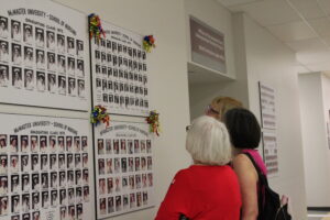 Three women standing in a hallway looking at a class picture hung up on the wall